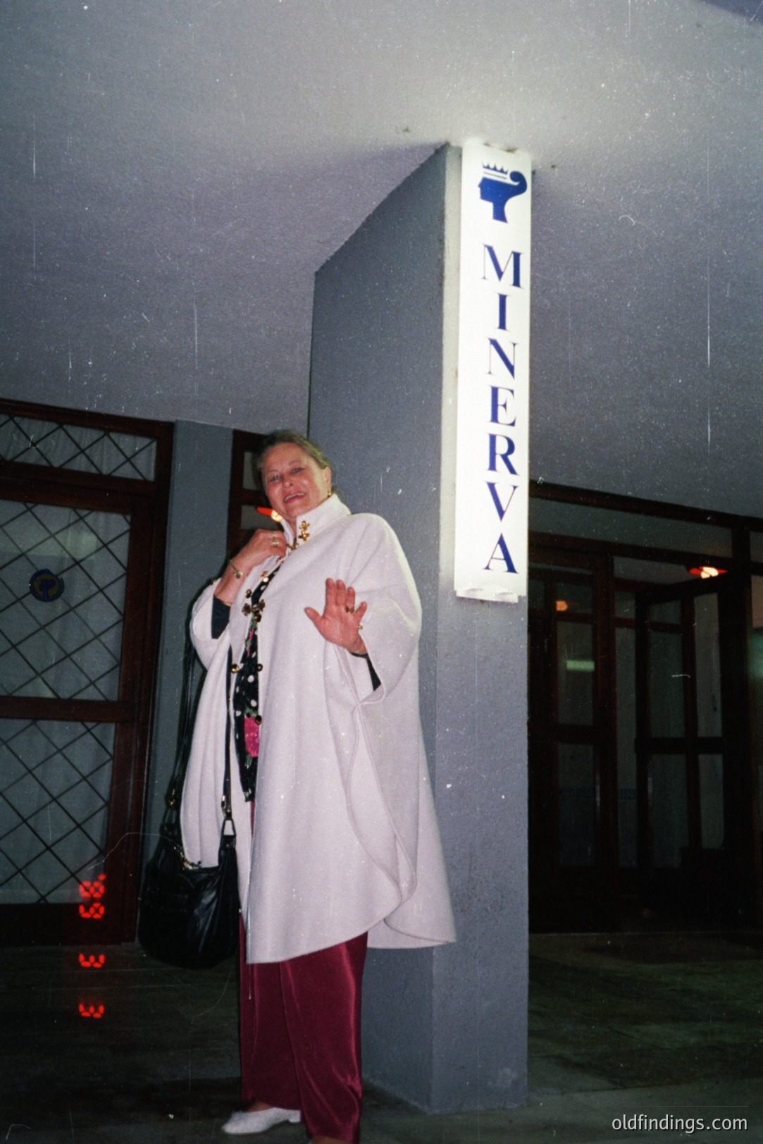 A woman in a white, flowing tunic and maroon pants stands outside a building with a “MINERVA” sign. Likely a hotel or resort, judging from the architectural details and signage. The photograph’s color palette and quality suggest a snapshot from the 1980s or 1990s. The woman holds a black bag, appearing relaxed and waving.