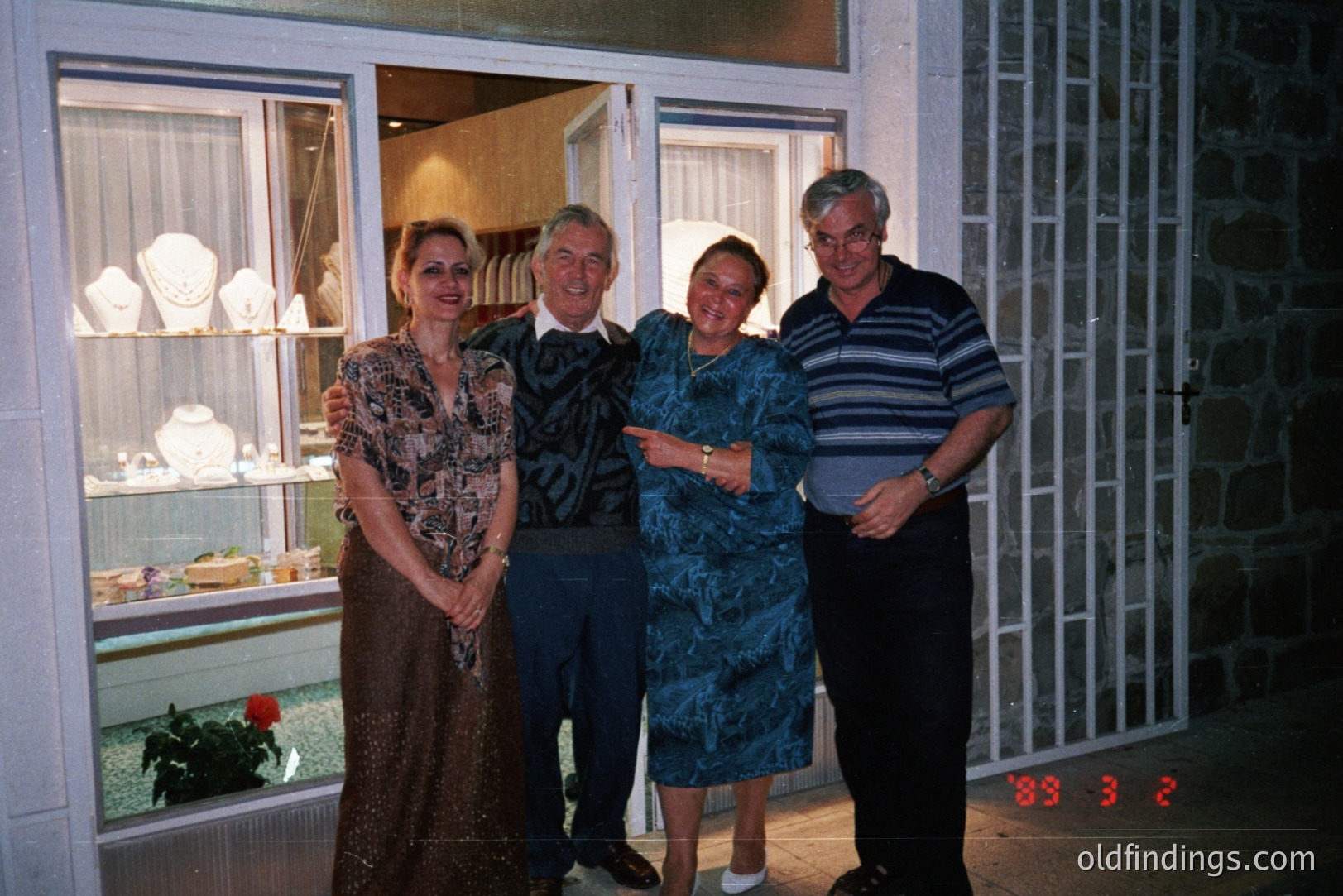 Four individuals pose outside a jewelry store; a woman in a floral dress, a man in a suit, another woman in a blue dress, and a man in a striped shirt. Store displays feature jewelry. Likely taken in the late 1980s based on the date stamp, possible vacation snapshot.