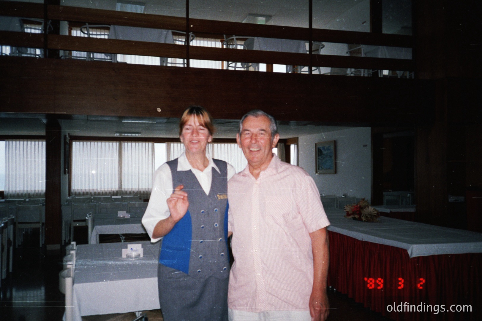 A smiling hotel employee in a dark blue uniform and a male guest pose for a photograph. The setting is a mid-century modern dining room, with large windows and elevated balcony. Dated stamp "88 3 2" suggests 1988. Likely a resort or hotel dining area.