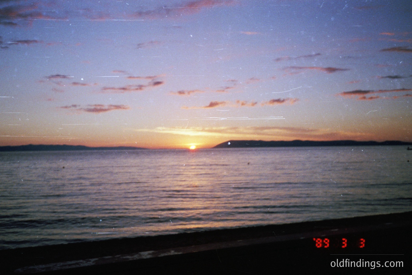 A tranquil seaside view captures a vibrant sunset over a calm body of water. The sky displays hues of orange, pink, and purple. A distant coastline silhouette is visible. The film grain and date code "89 3 3" suggest a late 1980s origin.