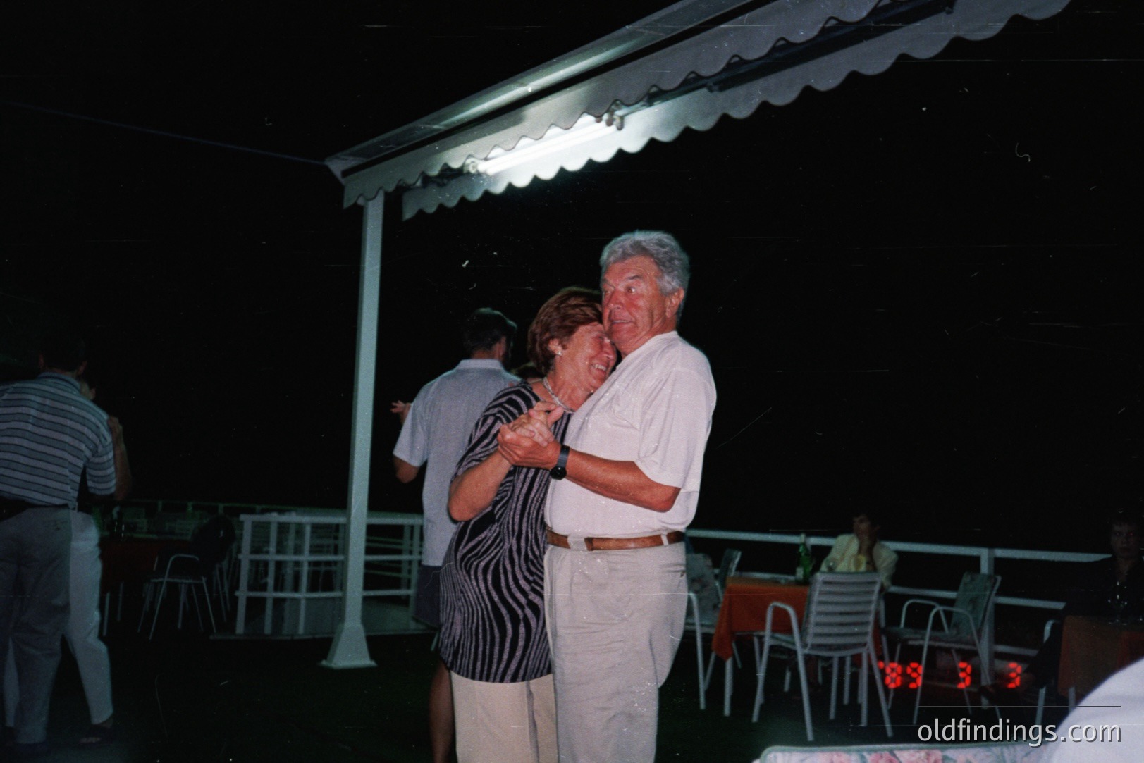 A couple dances closely beneath a covered patio, appearing joyful. The man wears a white suit; the woman, a patterned dark dress. Several guests and chairs are visible in the background, suggesting a party. Likely taken in the 1980s or 1990s, based on attire and film aesthetic.
