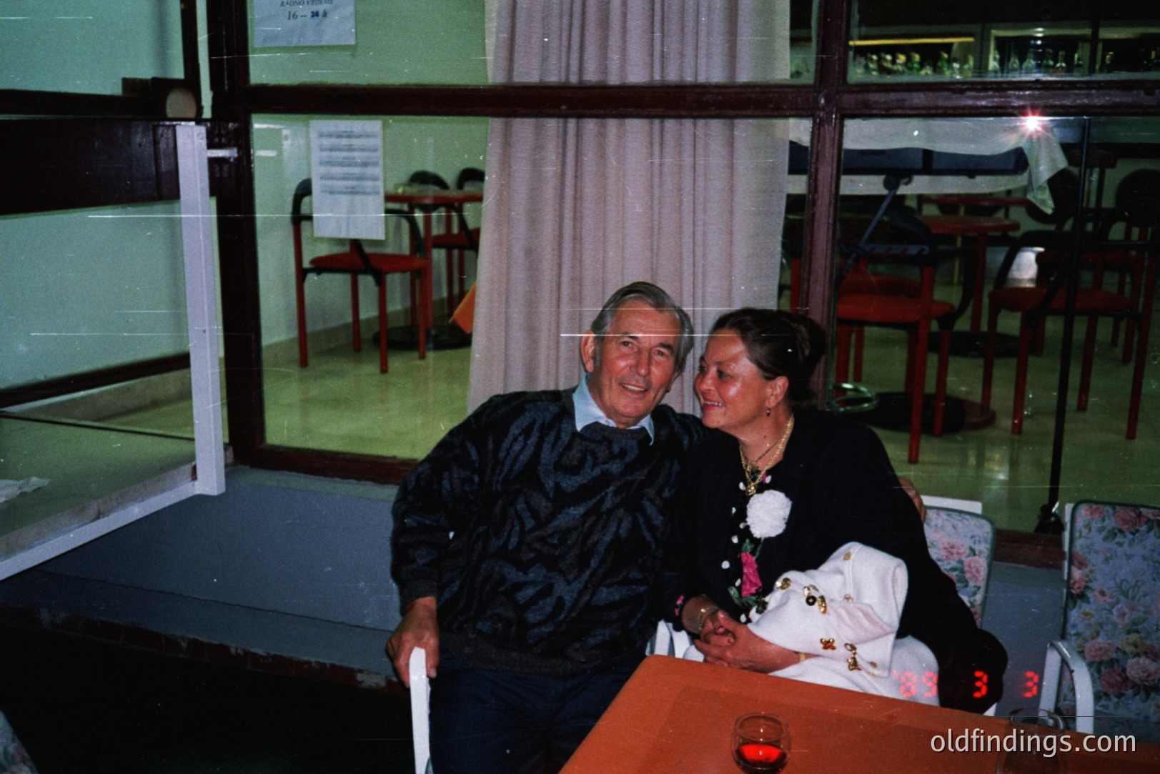 An elderly couple poses for a portrait in what appears to be a restaurant or banquet hall. Man wears a cable-knit sweater. Woman is in a dark dress and holding a decorative fan. Red-orange ambient lighting and visible film grain suggest a 1970s-80s origin.