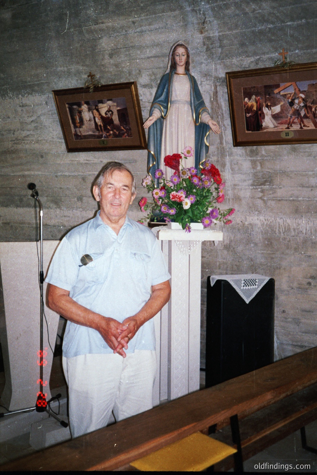 Man stands before a statue of the Virgin Mary in a stone-walled interior. White polo shirt, light-colored pants. Floral arrangement on a draped altar behind him. Simple, rustic decor suggests a small chapel. Likely a personal or community gathering space.