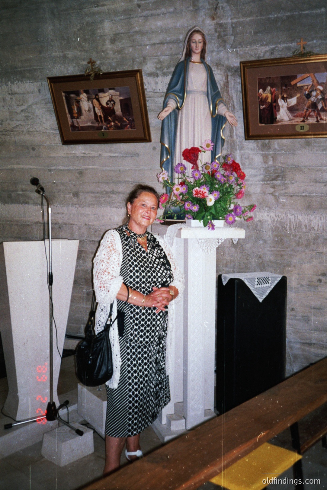 A woman stands in what appears to be a church or chapel. She wears a patterned, sleeveless dress with lace detailing. A statue of the Virgin Mary and floral arrangement stand behind her. The image exhibits a dated aesthetic, likely 1980s.