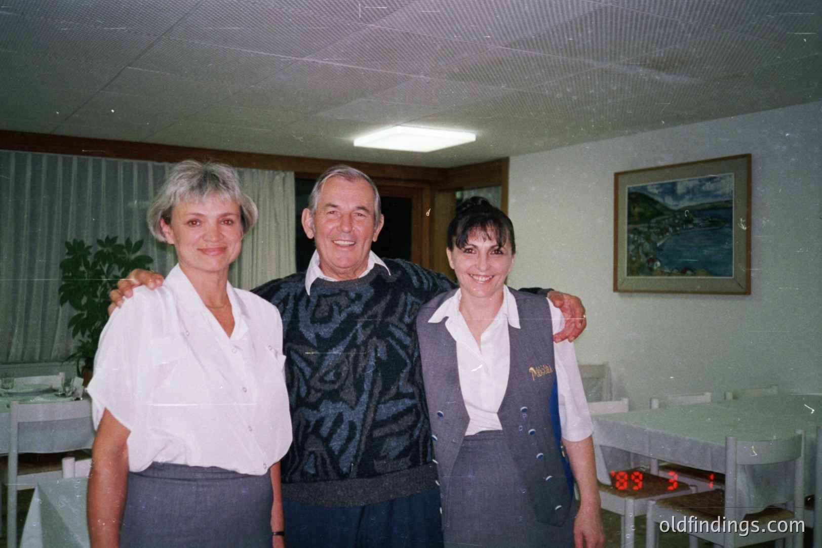 Three smiling individuals stand in a dining room setting. A man in a patterned sweater is flanked by a woman in a white shirt and grey skirt, and a woman in a server's uniform. Visible decor includes a seascape painting and potted plant. Likely a photograph from the 1980s, possibly a hospitality environment.