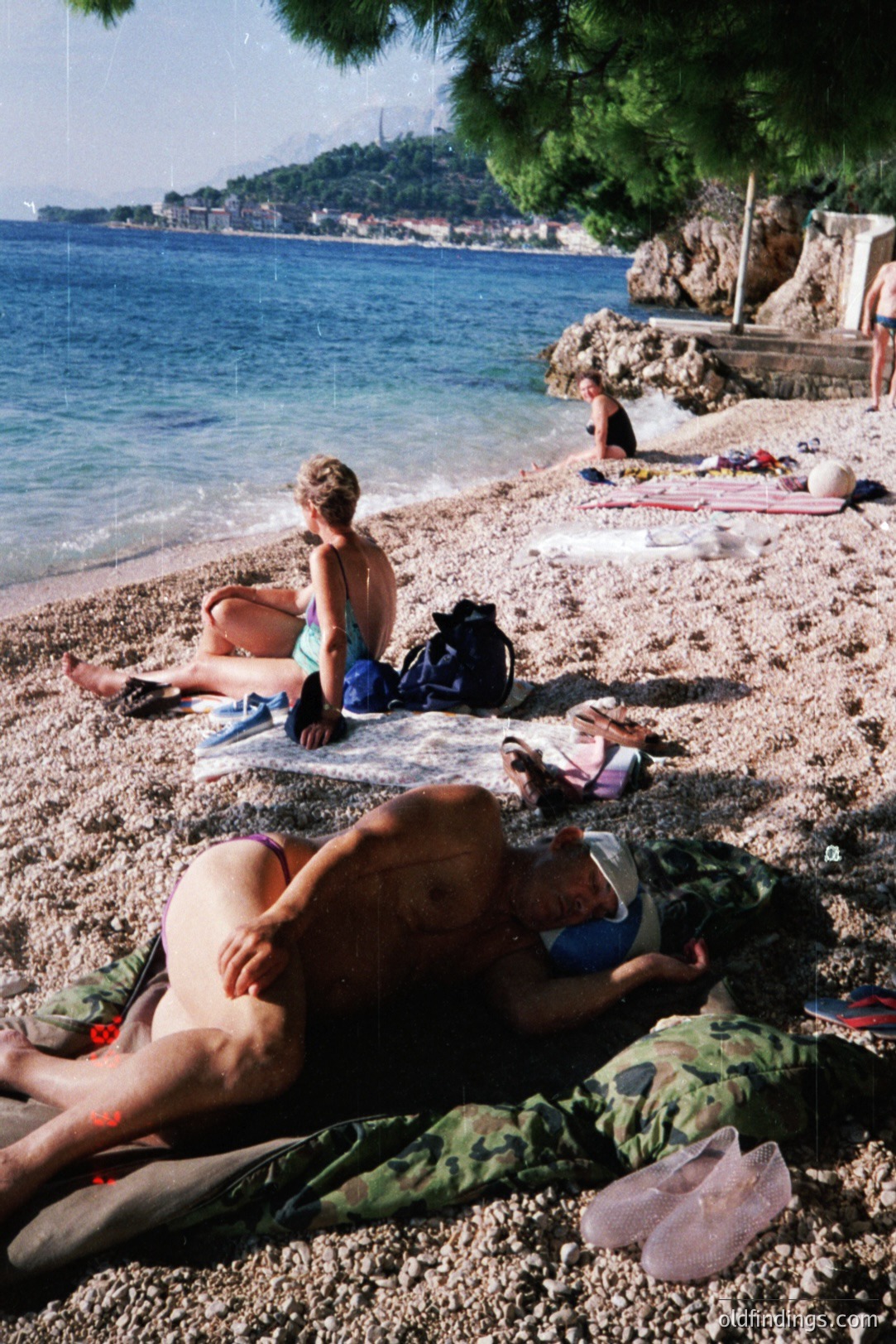Seaside view captures leisure time. Two women sunbathe on a pebble beach next to calm blue water. Buildings are visible along the hillside. Camo fabric and towels are near the subjects. Likely 1970s or 80s, possibly Eastern European coastline. Commercial value for travel or lifestyle stock.