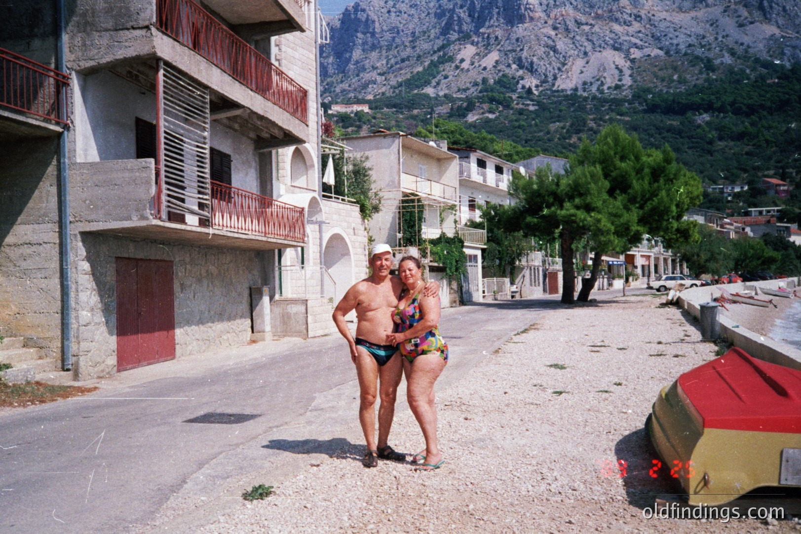 A couple stands on a coastal road, wearing swimwear, in front of modest apartment buildings. Lush, rocky mountains form a dramatic backdrop. Likely a seaside resort town, possibly in the Balkans. The grainy quality suggests a snapshot from the 1980s or 90s.