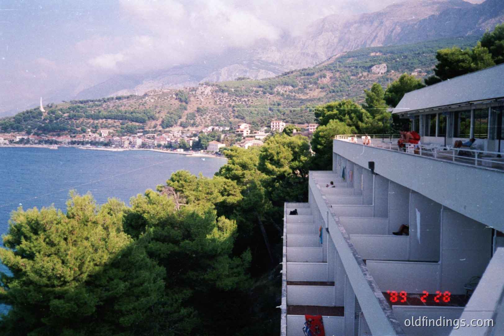 Seaside resort architecture, likely 1970s, overlooking a coastal town nestled between mountains and the Adriatic Sea. Concrete balconies extend from a multi-story building. Visible date stamp suggests 1988. Possible location: Yugoslavia (present-day Croatia/Montenegro).