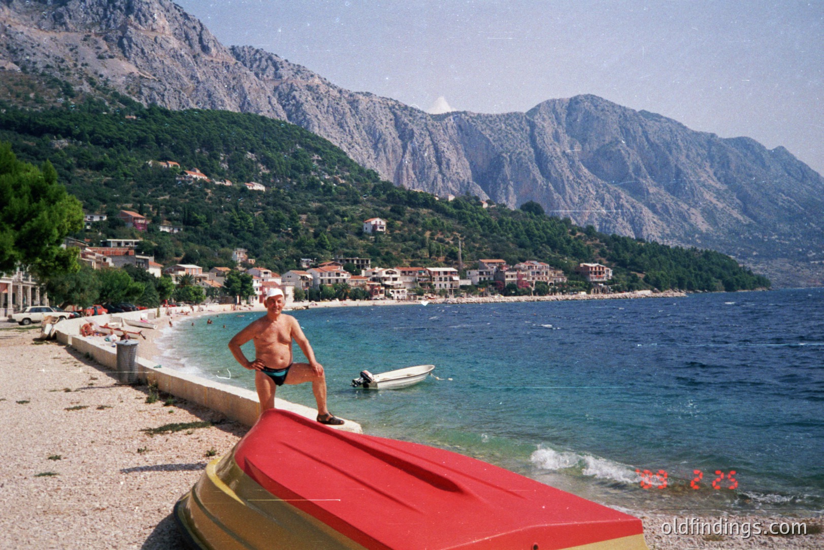 A man stands on a red and yellow kayak on a pebble beach, overlooking a coastal town nestled against steep, forested mountains. The scene captures a seaside village with traditional architecture and vibrant greenery. Likely taken in the 1990s. The landscape is striking, showcasing a European coastal environment.