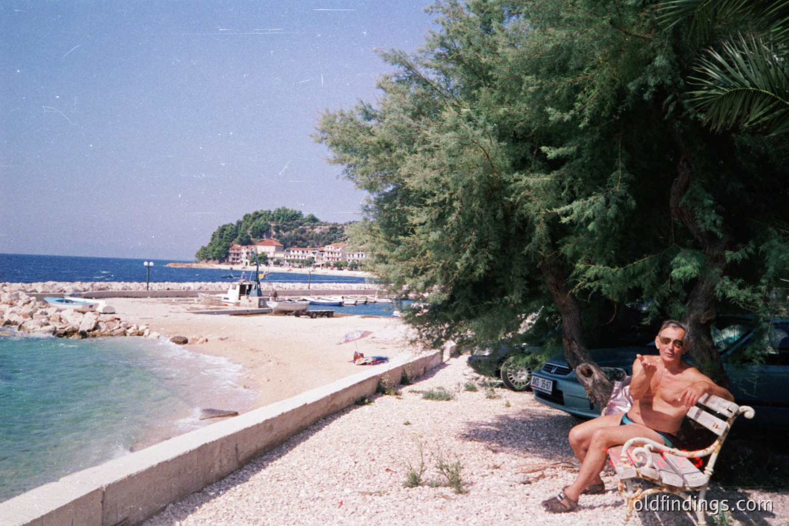 A man sits on a decorative iron bench, facing a pebble beach and turquoise sea. A dark green car is parked nearby, and lush greenery frames the view. Likely coastal Bulgaria; a small village is visible across the water. Appears to be a 1970s or 80s snapshot.