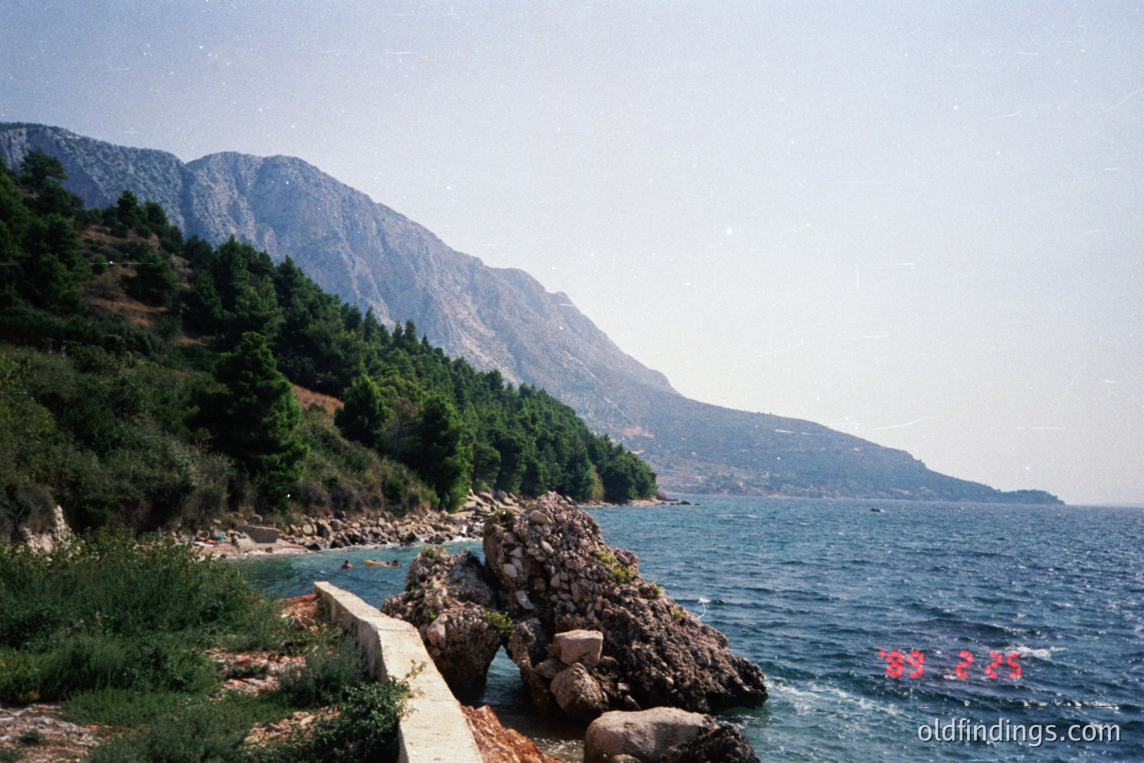 A coastal scene with rocky shoreline, dense pine forest on a steep hillside, and the Adriatic Sea. A concrete retaining wall is visible near the foreground. Date stamp indicates 1998. Possibly the coast of Montenegro or Albania. Likely a tourist destination.