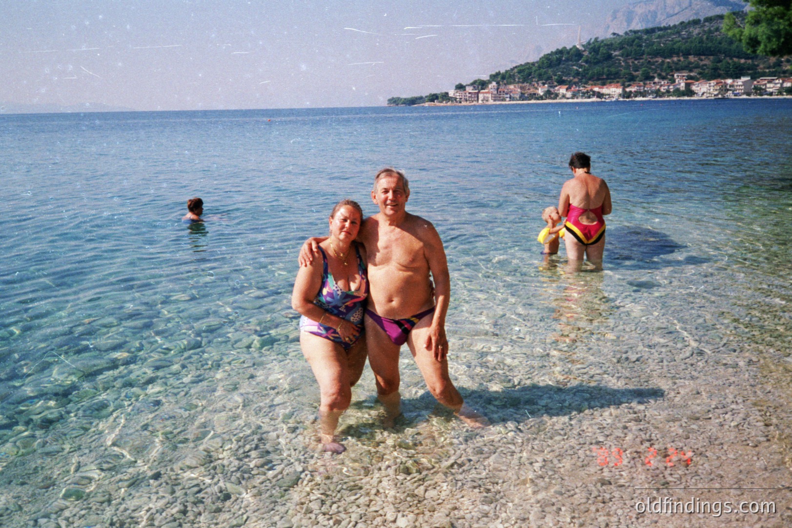 A middle-aged couple stands in shallow, clear water, seemingly on vacation. The man wears purple swim trunks, and the woman a blue one-piece. A woman and a child are wading further out. Background shows a coastal town nestled into a hillside. Likely 1980s-1990s snapshot.