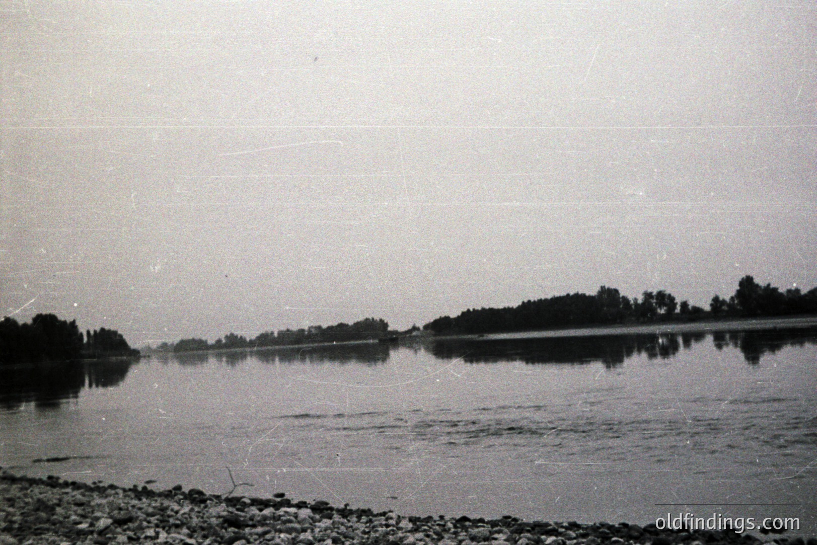 A wide, still body of water reflects the treeline on the opposite shore. A rocky foreground provides contrast. The scene appears tranquil, captured in black and white, with signs of aging on the film. Likely mid-20th century, potentially a landscape study. A valuable reference for graphic design or vintage aesthetic projects.