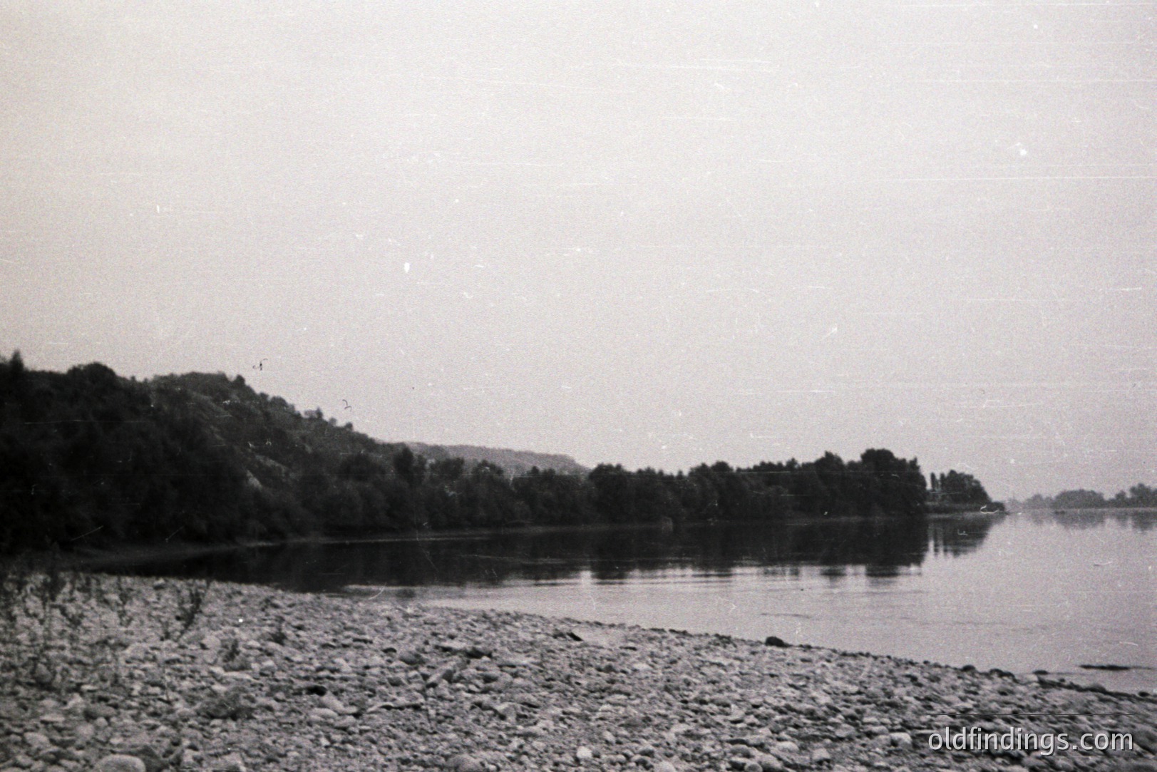 A monochrome, wide shot captures a rocky shoreline bordering a still body of water, likely a lake or calm sea. Lush, densely forested hills rise on the left, mirroring in the water. A distant structure sits on a promontory. Appears to be a lakeside scene. Likely mid-20th century, based on film grain and aesthetic.