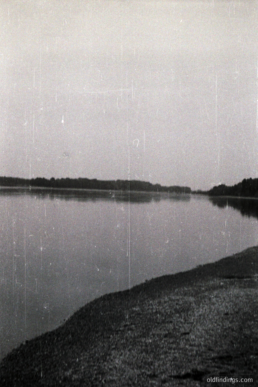 Still waters reflect a distant tree line. A rocky shoreline occupies the lower portion of the image, meeting a calm body of water under an overcast sky. Shows evidence of age/wear on photographic media. Likely mid-20th century amateur landscape. Could be suitable for vintage design projects.