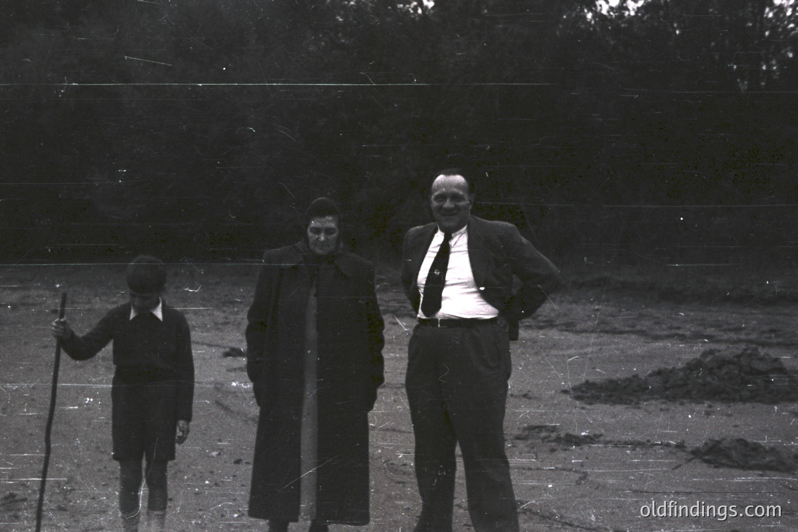 Three figures stand outdoors on a barren, sandy expanse. A young boy holds a stick, while an adult woman wears a long coat & headscarf. A man in a suit stands confidently, hands on hips. Likely a family portrait, possibly a vacation or rural setting. Estimated 1950s-1970s.