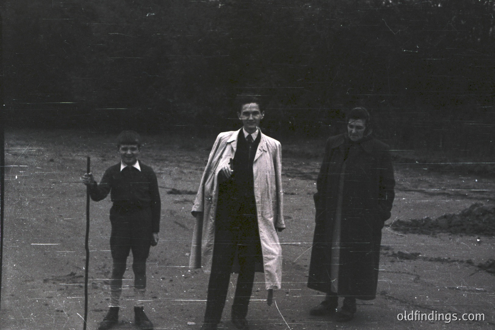 Three figures stand outdoors, appearing formally posed. The boy carries a wooden stick. The man in the middle wears a long coat and tie, likely 1960s or 70s attire. The woman's dark coat obscures details. The background suggests a wooded area.