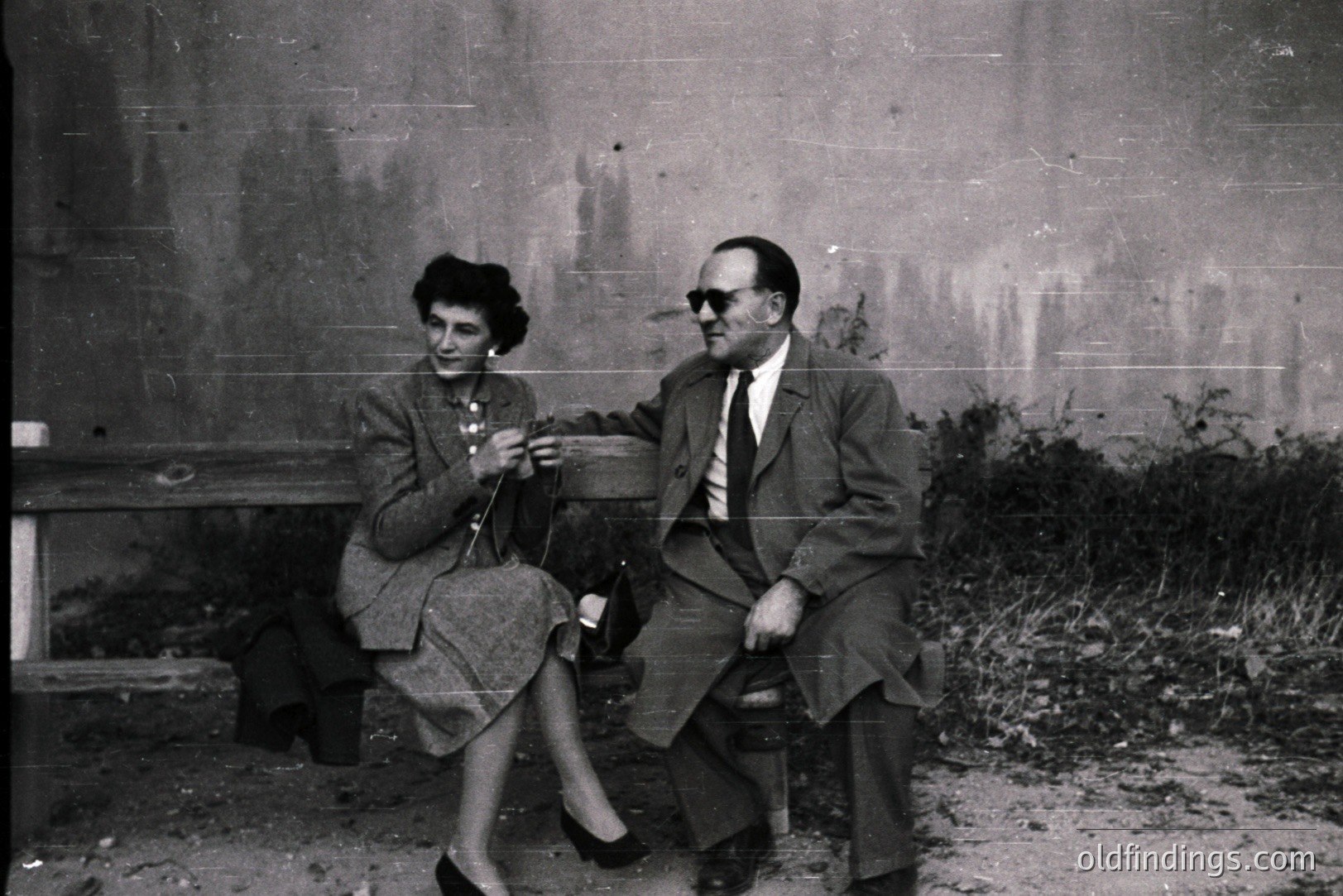 A formally dressed couple sits side-by-side on a weathered stone bench. The woman knits, while the man wears sunglasses. Visible architectural details suggest a European setting, possibly a courtyard or garden. Likely 1960s-1970s era. Potential stock photography value for period design.