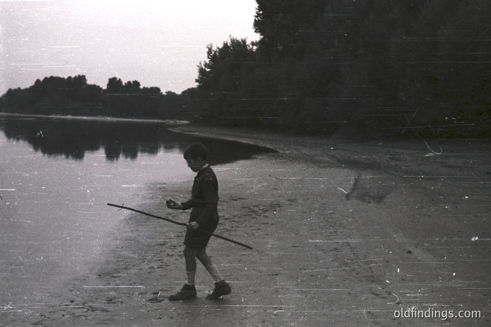 A young boy walks along a sandy shoreline, carrying a long stick over his shoulder. Trees line the opposite bank, reflected in still water. Likely a candid snapshot, possibly 1960s or 70s, evocative of childhood recreation by a riverbank. Potential for design/nostalgia themes.