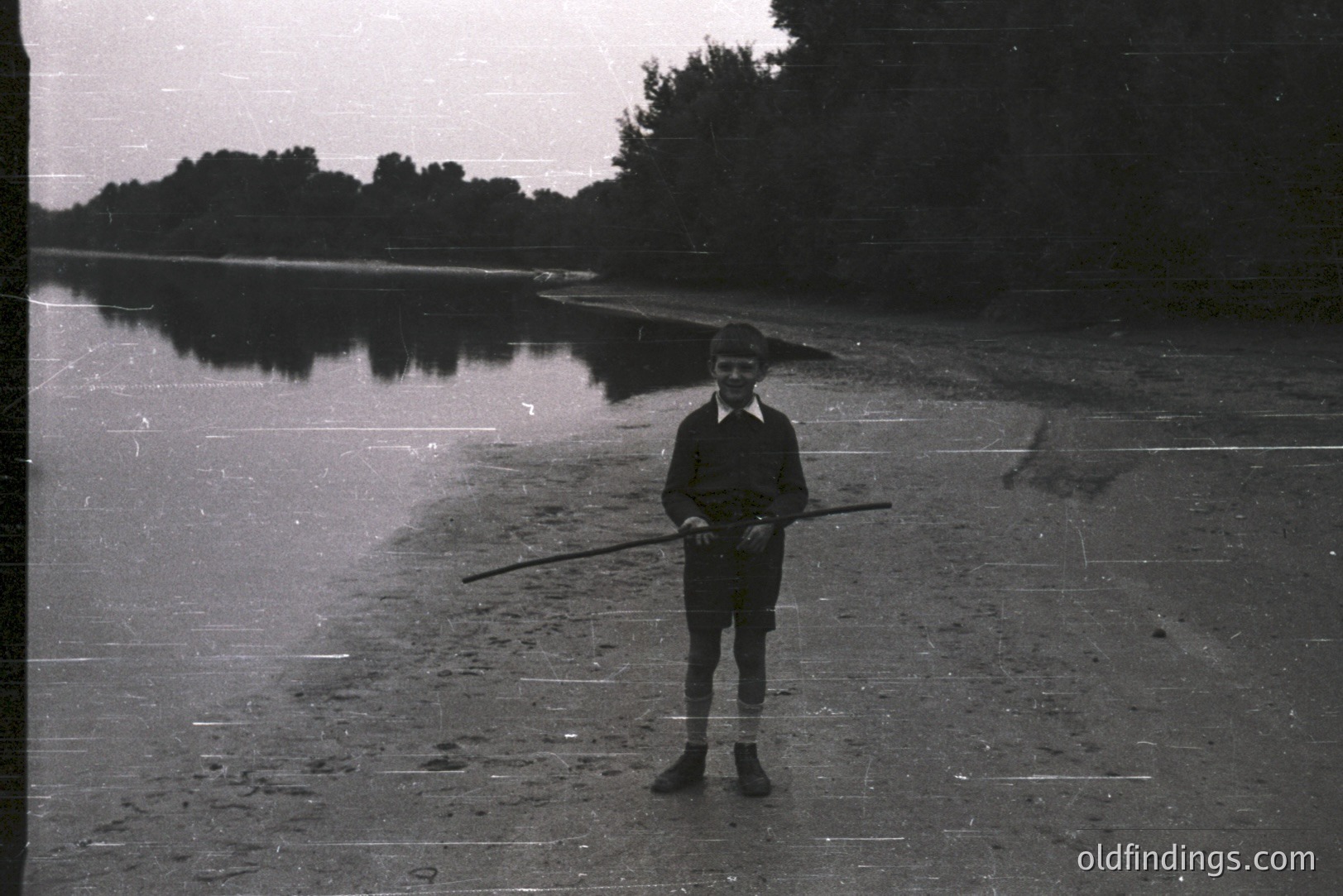 A young boy, wearing glasses and a dark collared shirt with short shorts, stands on a sandy shoreline holding a long, thin stick. A body of water reflects the treeline in the background. Likely a candid snapshot, indicative of mid-20th century casual dress. Potential stock photo value for vintage childhood themes.