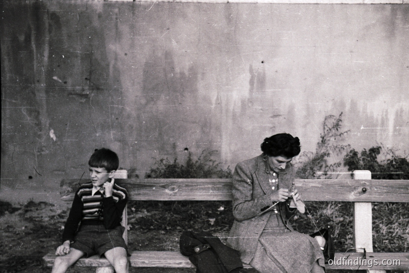 A young boy and woman share a weathered wooden bench. The boy, dressed in shorts and a striped shirt, appears thoughtful; the woman knits, facing away. Concrete wall and foliage backdrop suggest a public park or institutional setting. Likely 1950s-1970s. A valuable, candid moment for archival/historical use.