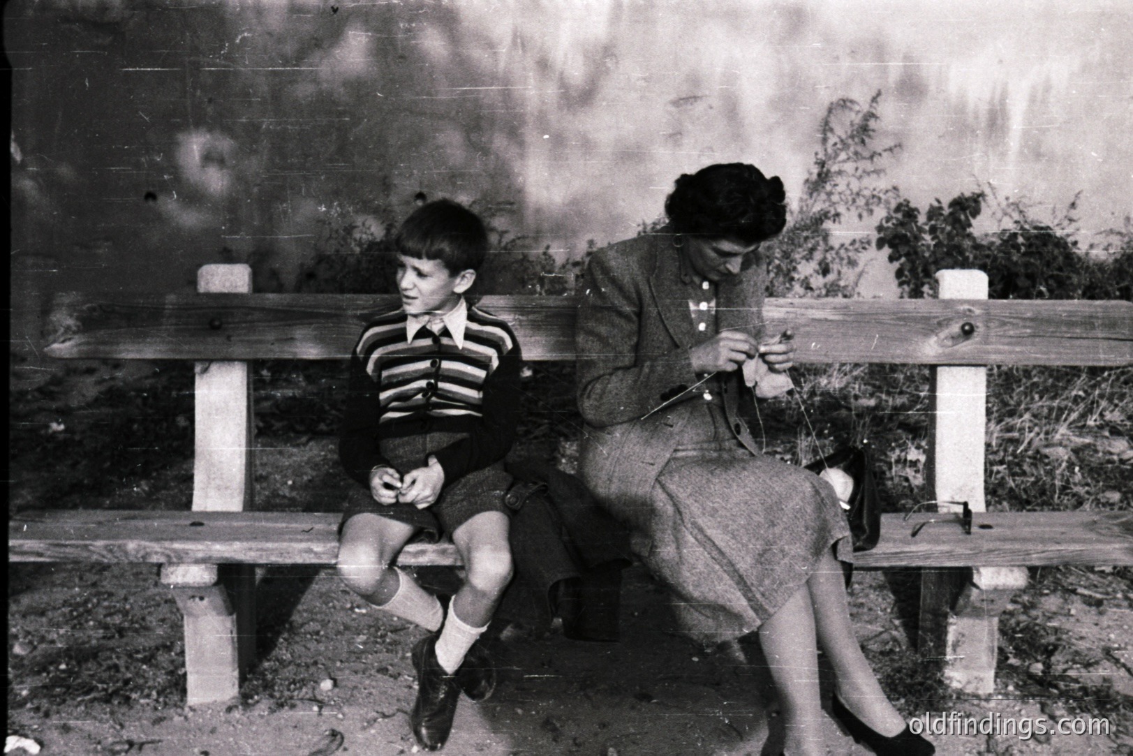 A young boy and woman are seated on a stone and wooden bench. The boy wears short trousers, knee socks, and buttoned sweater. The woman is knitting, dressed in a patterned dress and stockings. The background features foliage and a textured stone wall. Likely 1950s, possibly European. A quiet, domestic moment.