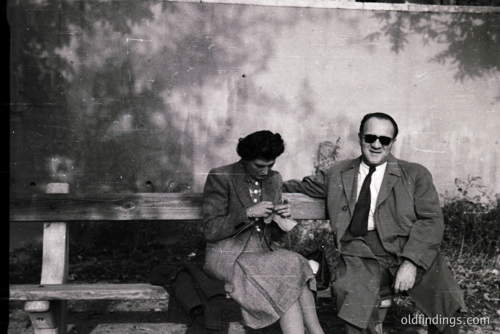 A man and woman are seated on a weathered stone bench, set against a textured concrete wall with foliage shadows. The woman knits, while the man wears a suit and sunglasses. Likely a mid-century snapshot, possibly of a vacation or leisure time. Simple composition, strong architectural backdrop.