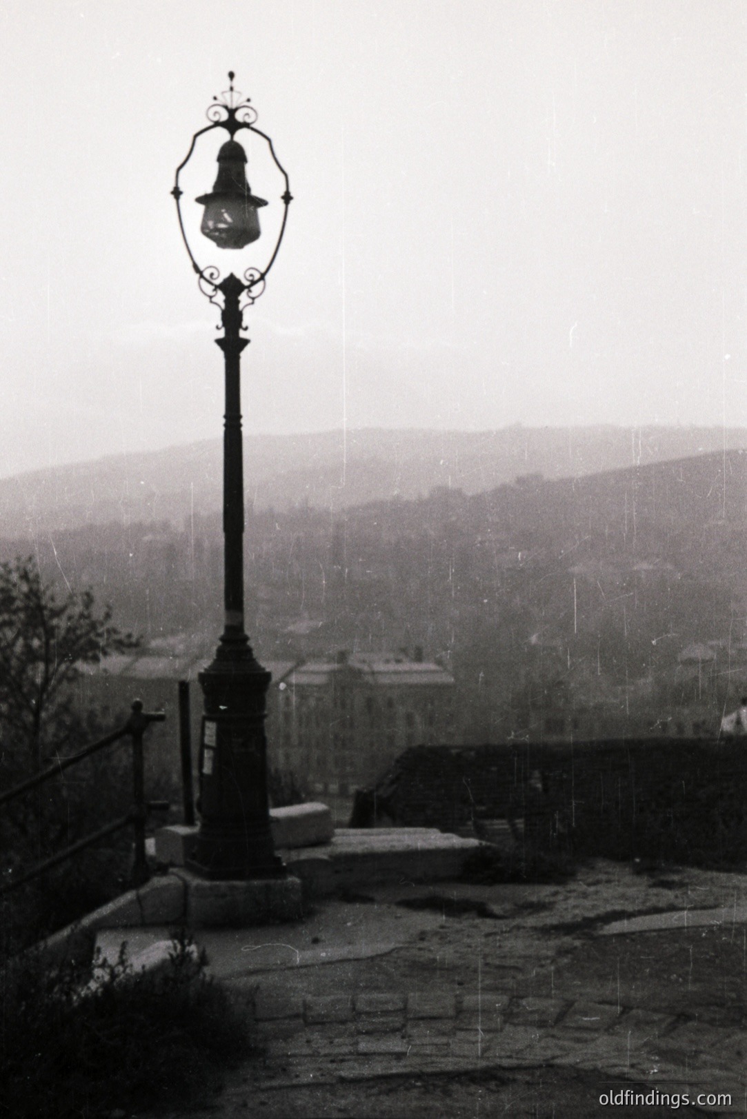A wrought iron lamppost stands prominently on a stone ledge overlooking a city panorama, likely a European coastal town. The scene suggests a slightly misty, overcast day. Architecture hints at a historical or early 20th-century setting. A handrail guides a stepped path leading down.