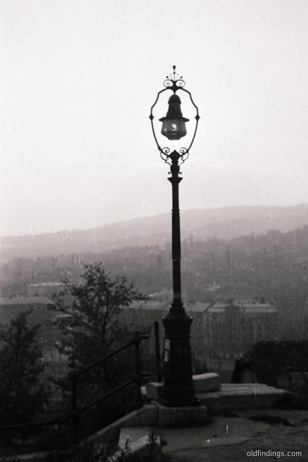 Ornate, wrought-iron lamppost overlooking a tiered townscape, likely a European coastal city. Visible steps and ornate railings suggest a formal garden setting. The photograph’s texture hints at early photographic processes (e.g., albumen print). Likely dating from the late 1800s. Excellent reference for architectural details.