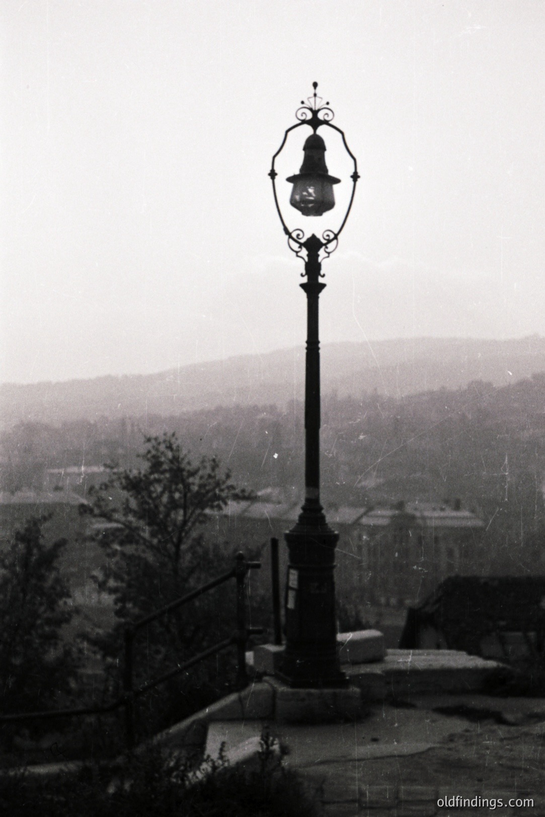 Ornate cast-iron lamppost stands overlooking a town nestled within a valley. Stairway & partial foliage visible. The misty atmosphere suggests a damp climate. Architectural style hints at early 20th century design. Potential stock photography reference.