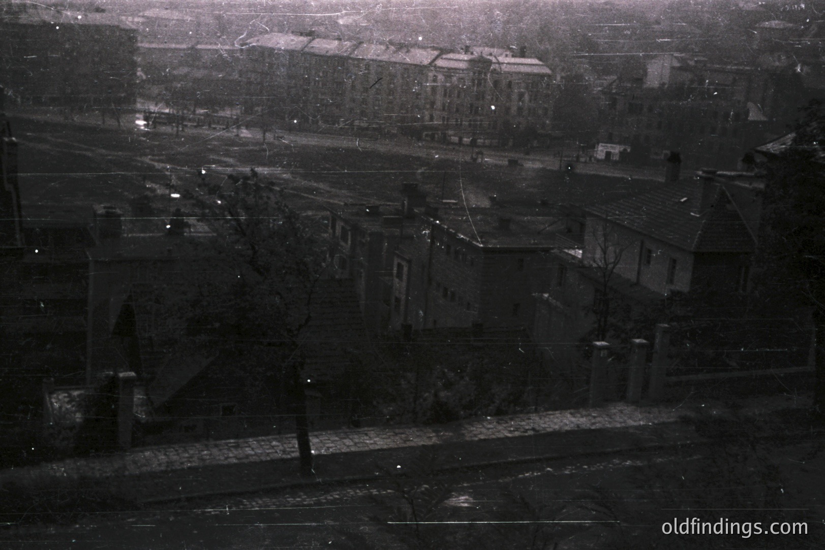 High-angle, grayscale view of a residential neighborhood, likely post-war era. Buildings feature steep roofs and varied architectural details. Paved street descends toward a wide, open space. Shows urban planning & infrastructure development. Potential use for historical context or architectural reference.