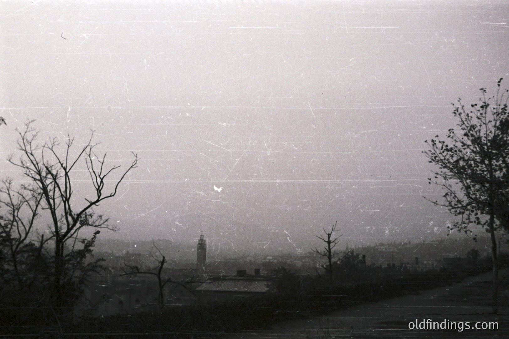 Atmospheric, grainy black and white landscape depicts a town viewed through bare branches. A tall tower is central; rooftops and buildings suggest a small European settlement. The image exhibits signs of age and possible storage damage. Likely 1960s-1970s era.