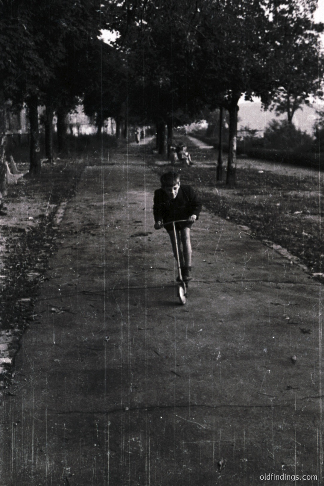 A young boy scooters down a tree-lined path, likely in a park. Details suggest a mid-20th century (1950s-1970s) aesthetic. Visible grain & scratches add to vintage feel. Other figures are seen in background. A study in motion.