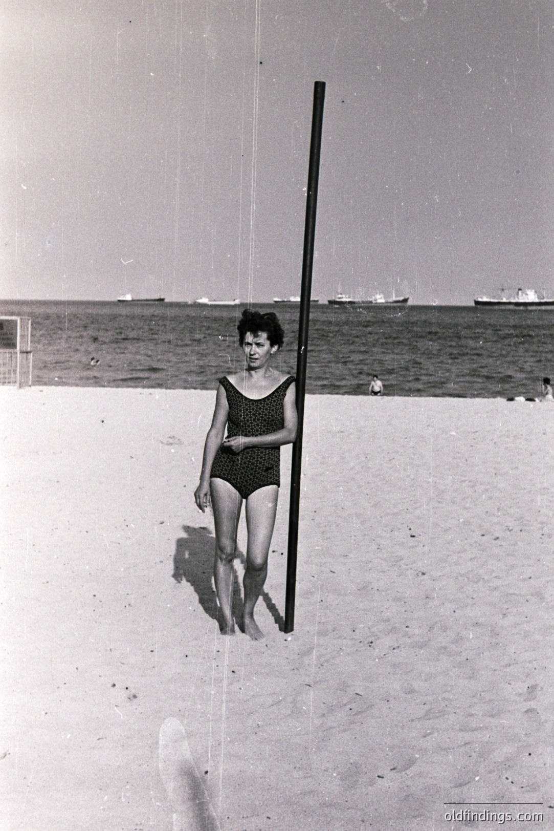 Black and white image shows a woman in a vintage swimsuit standing on a sandy beach with a long, dark pole positioned vertically beside her. A seascape and distant boats form the backdrop. Likely 1960s-1970s, possibly a seaside resort. The composition suggests a staged portrait or casual snapshot. Commercial potential for vintage advertising or fashion design references.