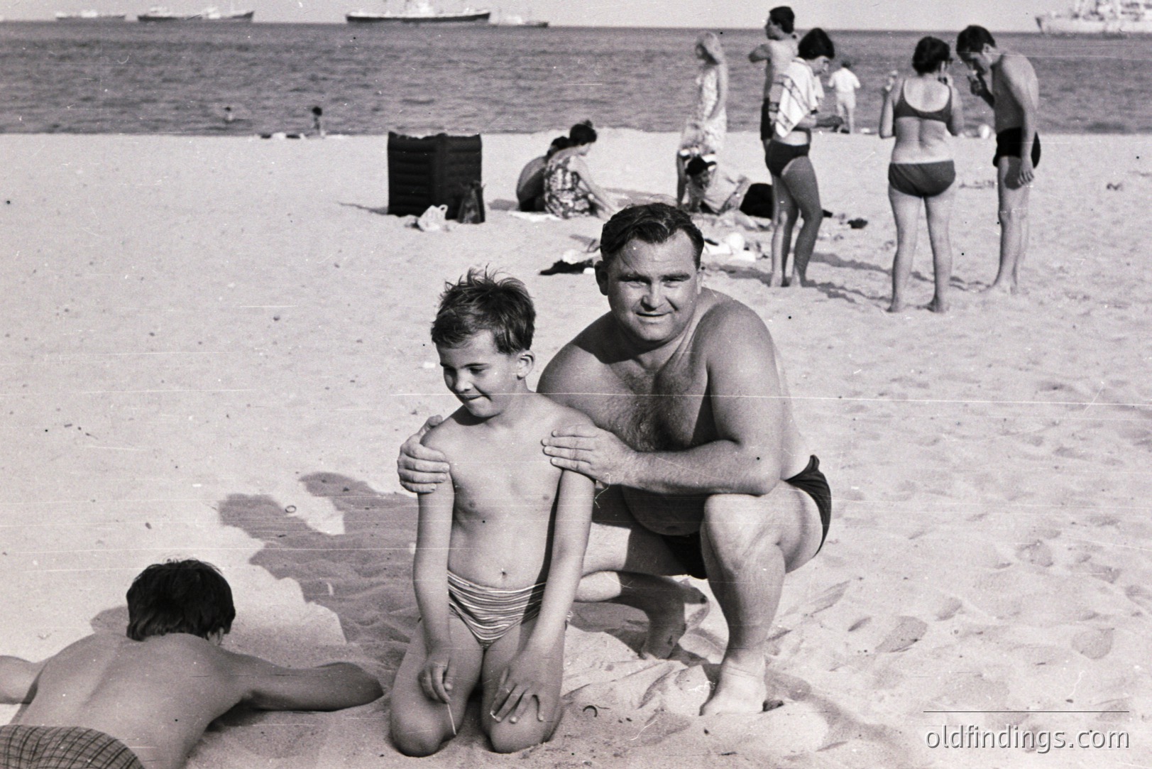 A father and young son are centrally posed on a sandy beach, with other beachgoers visible in the blurred background. The man wears black swim trunks; the boy, patterned briefs. A relaxed, candid moment likely captured in the 1960s or 70s. A ship is visible at sea.