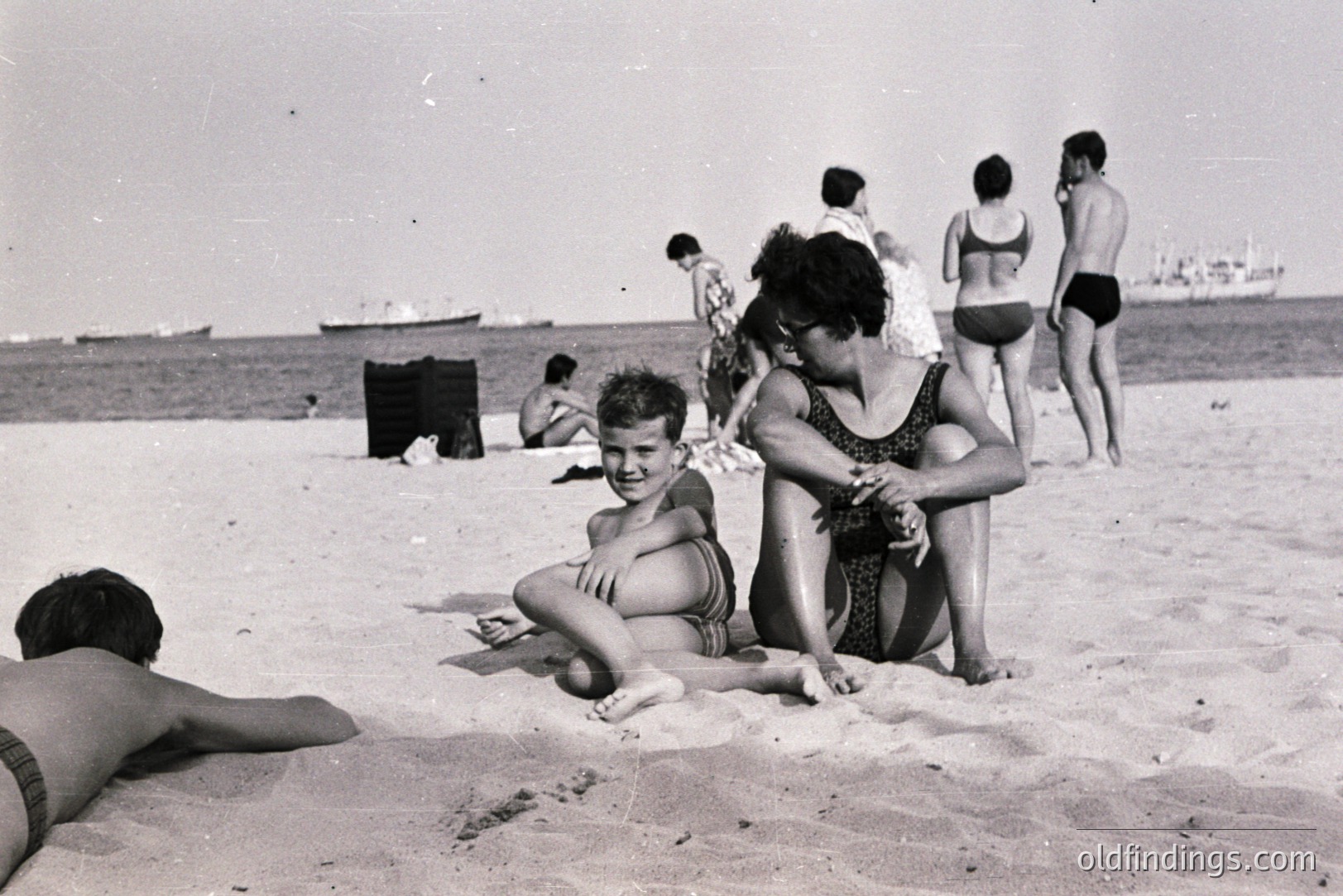 A candid moment on a beach, likely in the 1960s or 70s. A young boy and woman sit close on the sand, facing the camera. Several other figures are in the background, enjoying the seaside. Visible ships are on the horizon. Classic swimwear styles and grainy texture suggest the era.