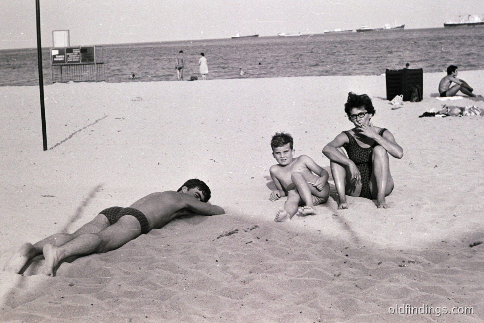 A candid beach scene captures three individuals enjoying a sunny day. A young man sunbathes, while a boy sits cross-legged and a woman in a swimsuit observes. Shipping vessels are visible on the horizon. Likely 1960s/70s coastal leisure.