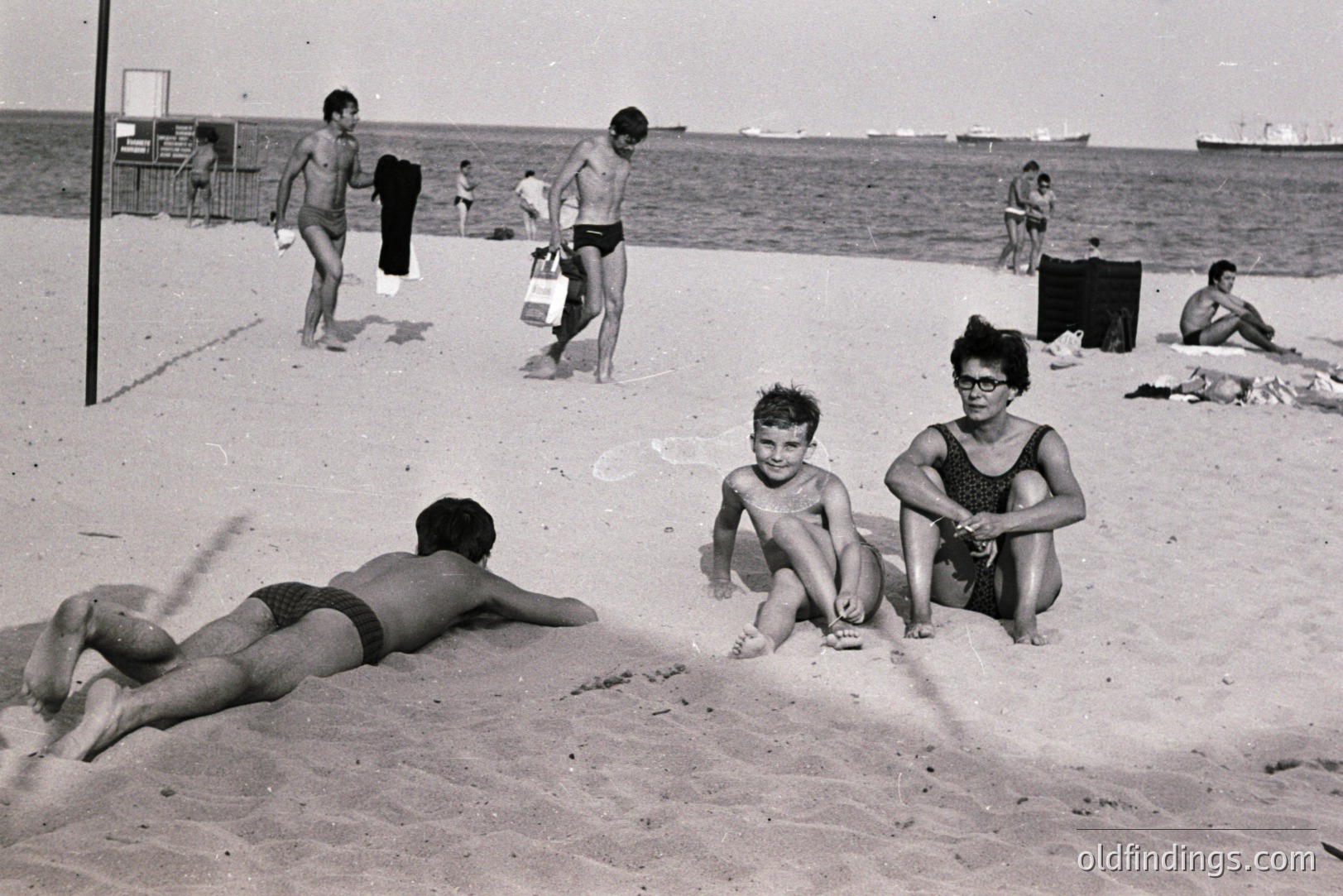 Black and white image depicting a beach scene, likely mid-1960s. Several people are visible, including boys playing, sunbathers, and a woman with glasses seated on the sand. A large ship is visible on the horizon. The grainy quality suggests vintage print.