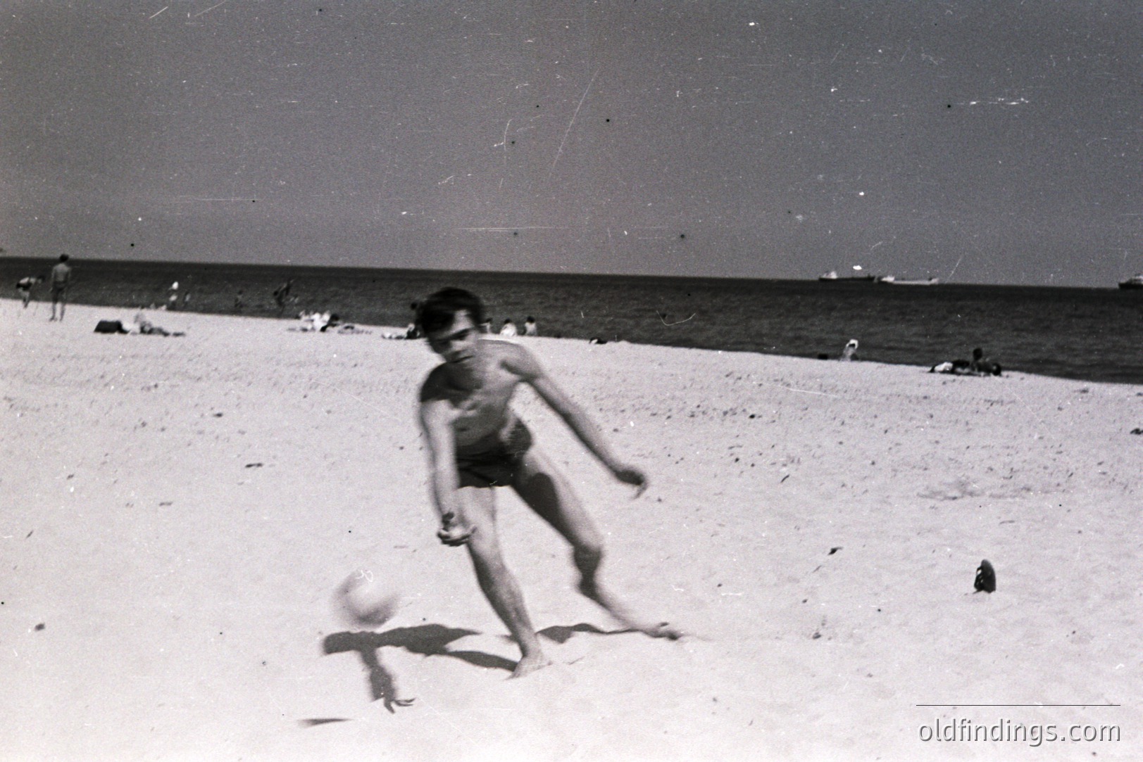 A barefoot boy kicks a ball on a sandy beach. Several other figures are visible in the background, enjoying a sunny day by the sea. Likely a seaside resort, potentially in Eastern Europe. Appears to be a snapshot from the 1960s-1970s. A candid moment capturing leisure and recreation.