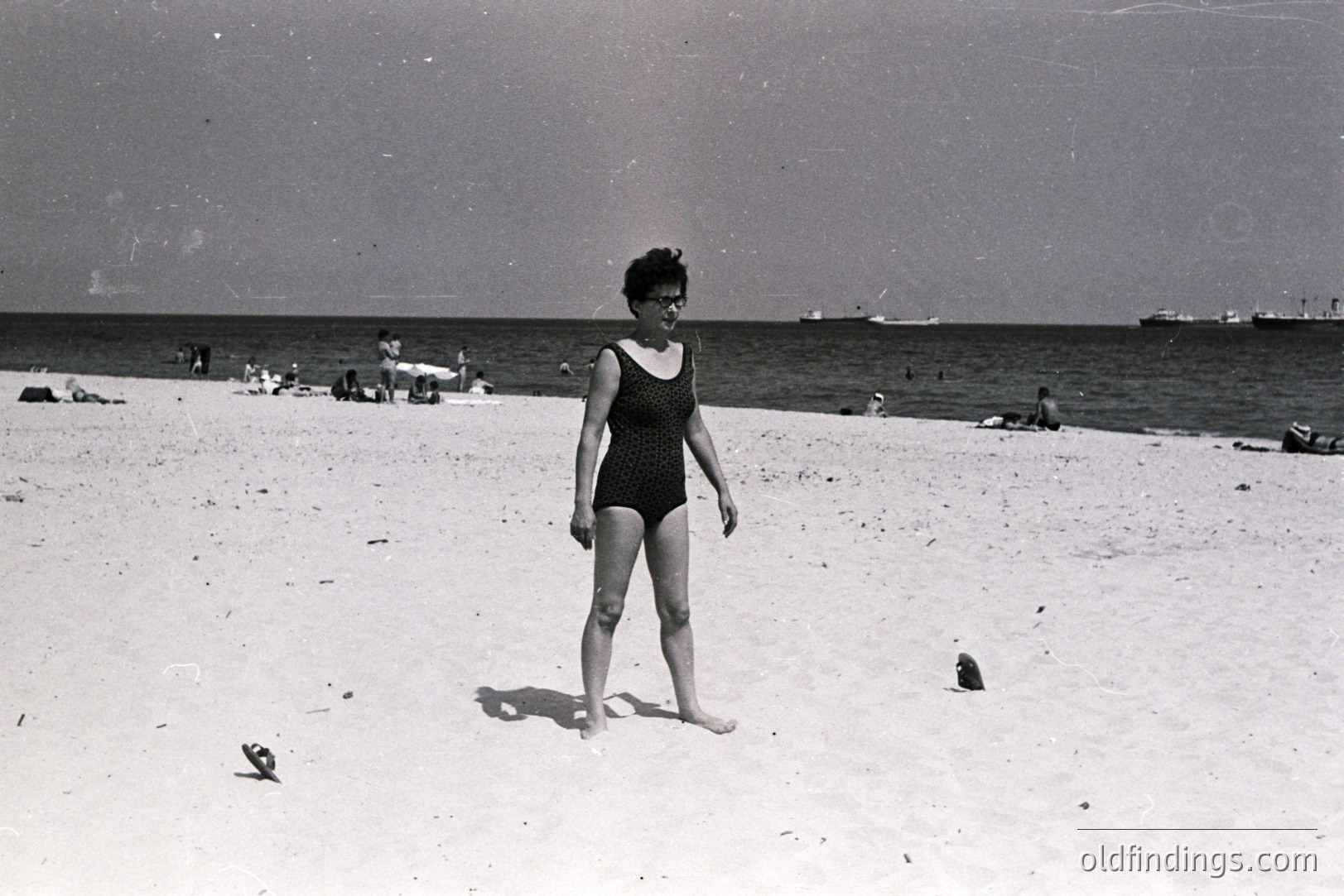 Black and white image captures a woman on a sandy beach, likely in Bulgaria, circa 1960s. She wears a vintage one-piece swimsuit and short, styled hair. Several figures and a ship are visible in the background. Possibly a seaside resort vacation snapshot.