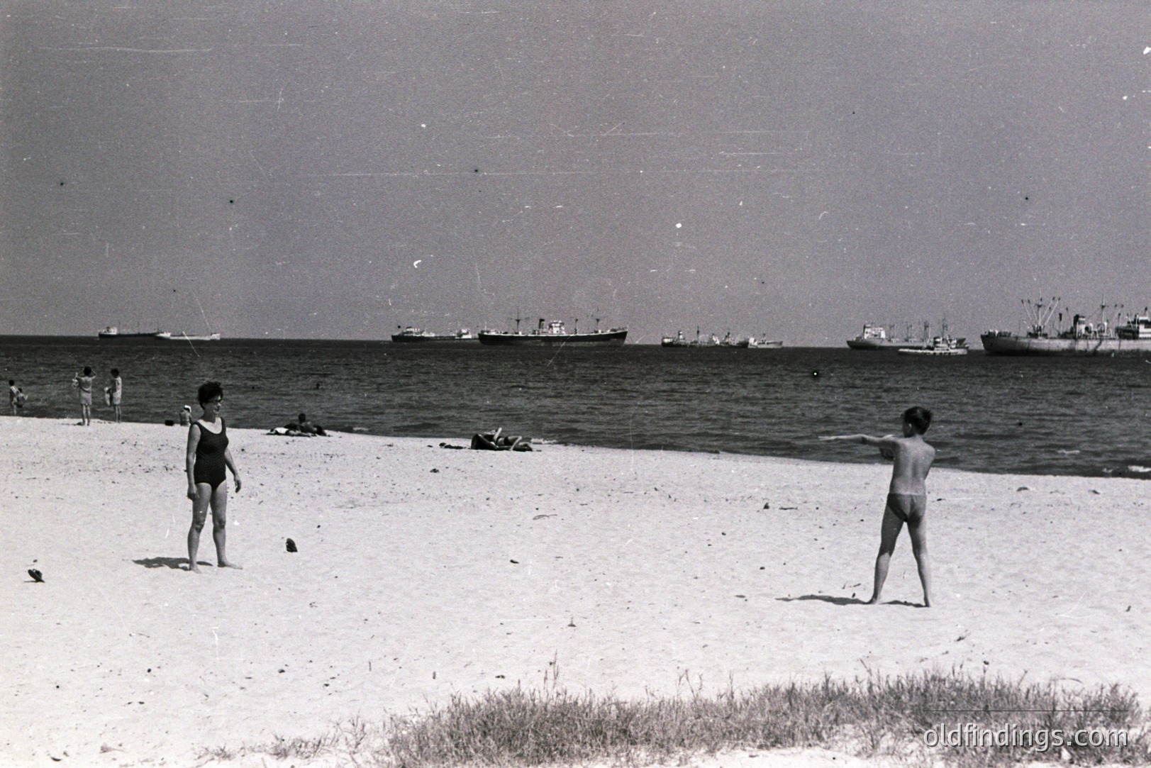 Black and white image depicting a seaside scene. Two children appear on a sandy beach, facing each other. Several cargo ships are visible in the background, indicating a port location. Likely taken in the 1960s or 70s, showcasing a moment of leisure.