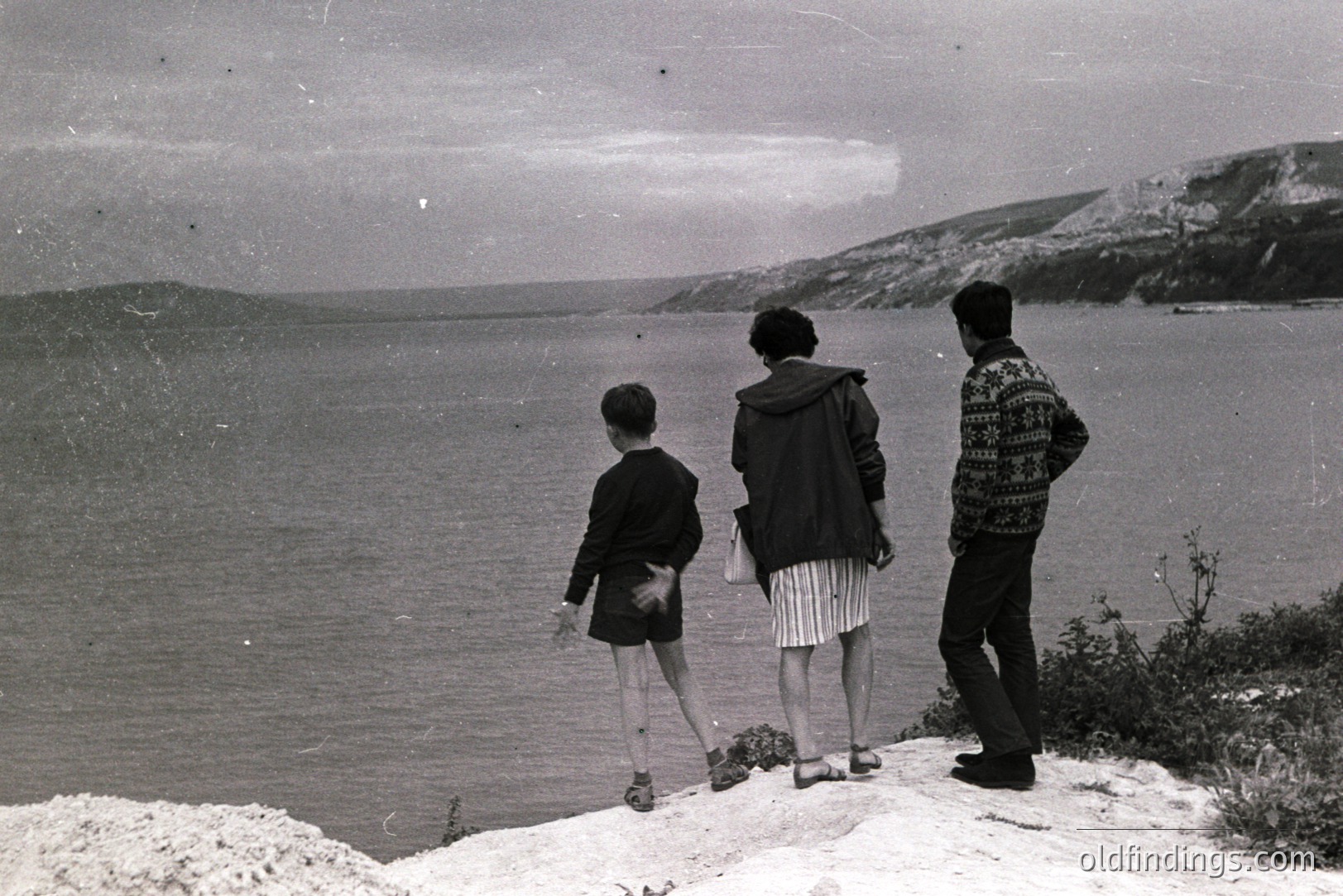 A young boy, a woman, and a man stand on a rocky coastal promontory gazing out over the expansive sea. The man wears a patterned sweater and the woman a dark jacket. Minimalist composition; stark, documentary style. Likely a family vacation photo. Estimated 1960s-1970s.