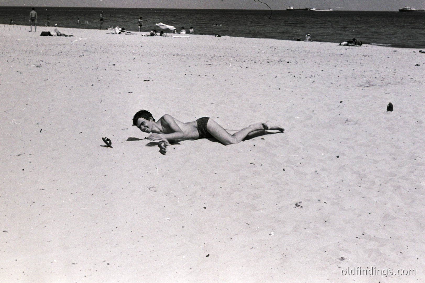 A young man in swimming trunks lies face-down on a sandy beach, arms outstretched. Distant figures populate the shoreline and ocean. Sparse vegetation and a clear sky complete the scene. Likely a candid moment from leisure time, potentially 1960s-1970s. A single bird is visible near the subject.