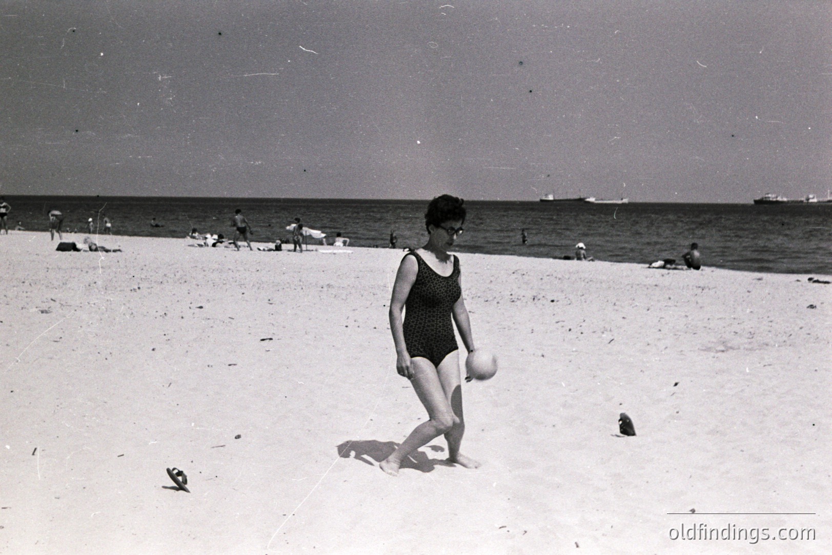 A woman in a black one-piece swimsuit walks along a sandy beach, clutching a beach ball. Several figures are visible in the background, enjoying the seaside. Likely a 1960s vacation scene. Grainy black and white image with visible film artifacts.