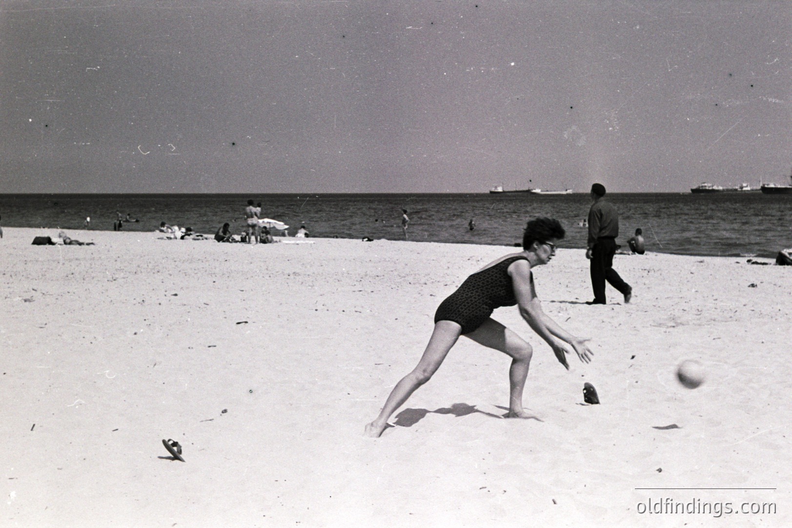 A woman in a one-piece swimsuit leaps forward to catch a ball on a sandy beach. Background shows other beachgoers and ships at sea. Likely a seaside resort vacation scene, possibly 1960s. Simple, graphic composition. A candid moment of leisure.