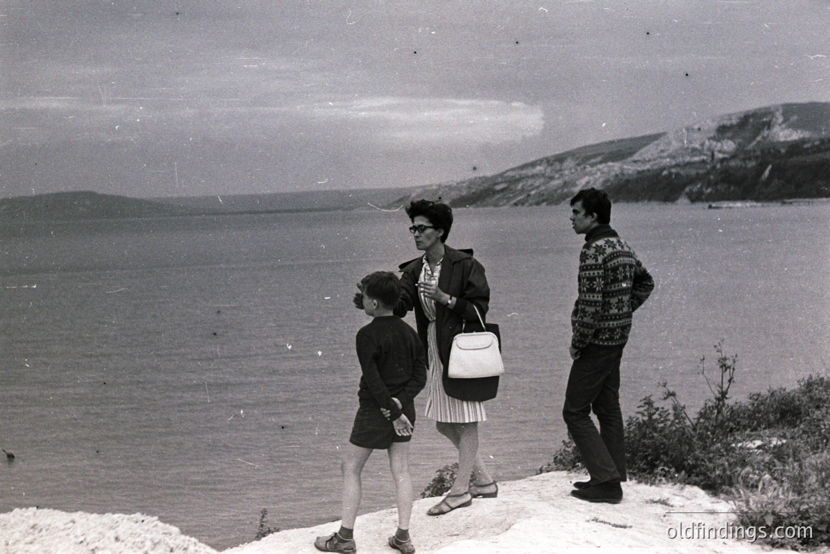 A family - woman, man, and child - stand overlooking a vast seascape. The woman wears a dress and carries a handbag; the man, a patterned sweater. The backdrop features a rugged coastline. Likely a seaside holiday scene, circa 1960s-70s.