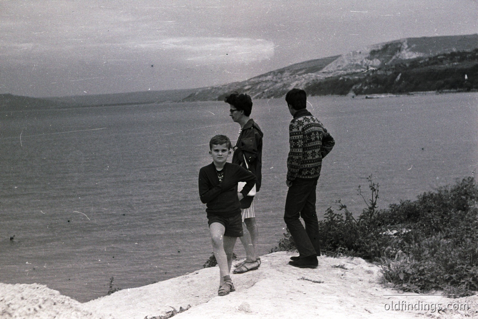 A young boy runs along a rocky clifftop overlooking a wide expanse of sea. Two other figures, one in a jacket and one in a patterned sweater, stand observing. Likely a coastal scene, possibly Britain, captured in a grainy, vintage aesthetic. Appears to be a candid snapshot from the 1960s or 70s.