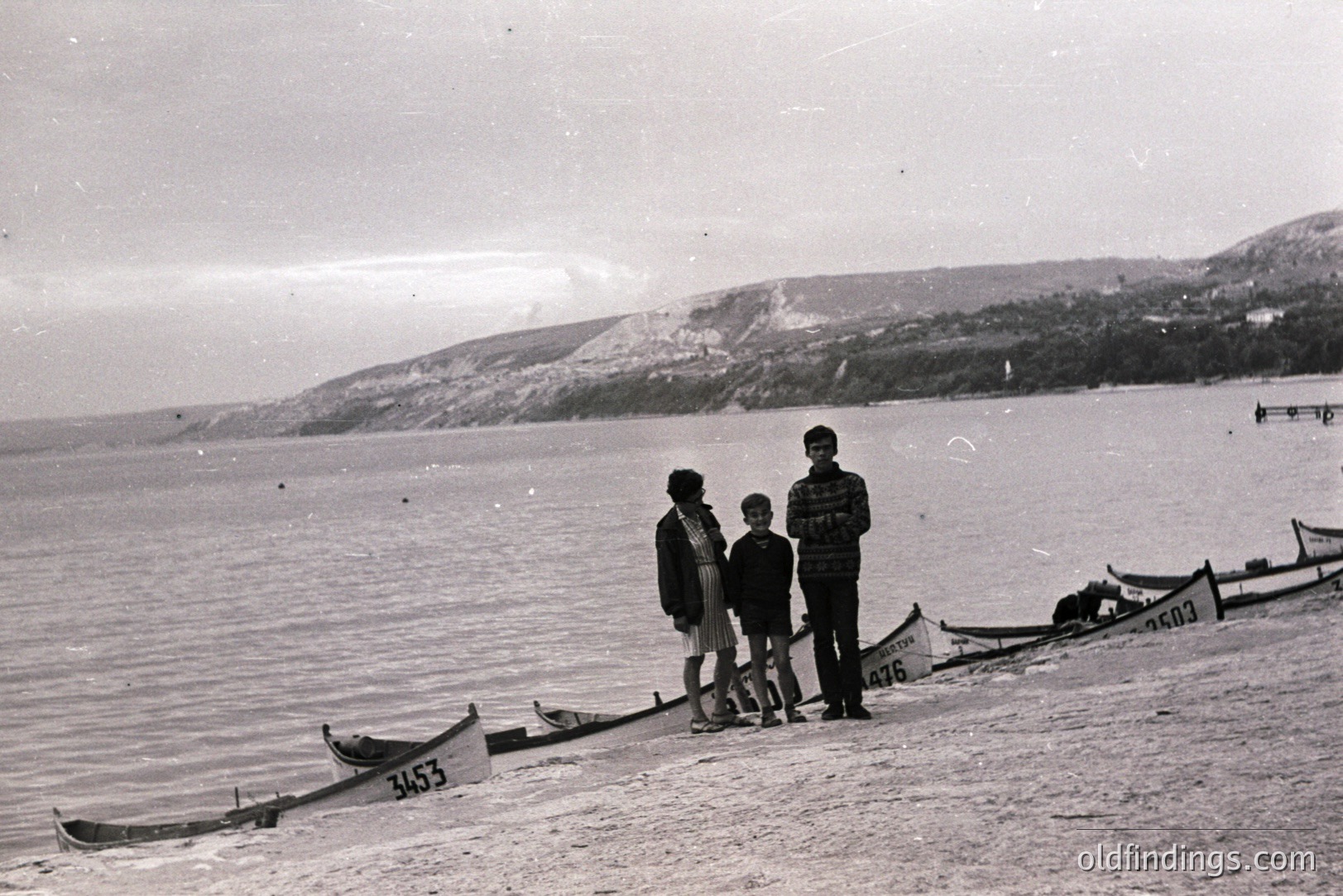 Seaside scene featuring three figures—two boys and a man—standing among rowboats on a sandy beach. A hillside with sparse vegetation rises in the background. Appears to be a coastal town, possibly Eastern Europe, 1960s-1970s. Rowboats are numbered, likely for a regatta.