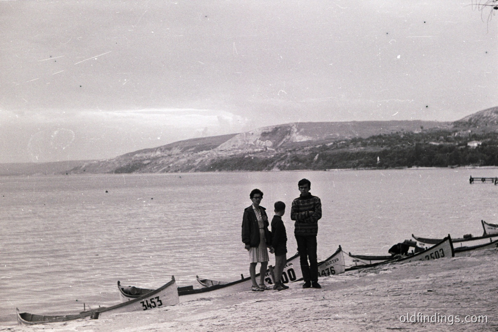 Three figures stand on a beach beside an array of small, numbered boats, likely canoes. The background features a gently sloping shoreline and distant, rocky hills. Likely 1960s, possibly a coastal resort setting. Image has vintage film grain and wear. Could be valuable for historical or design applications.