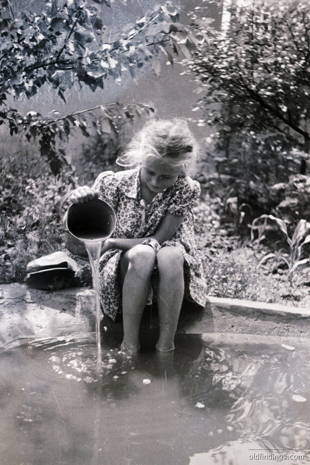 A young girl in a patterned dress sits at the edge of a stone basin, carefully pouring water from a bucket. Lush foliage surrounds the scene, hinting at a garden setting. Likely a mid-century snapshot, capturing a simple, everyday moment. The grainy texture is characteristic of older film stock.